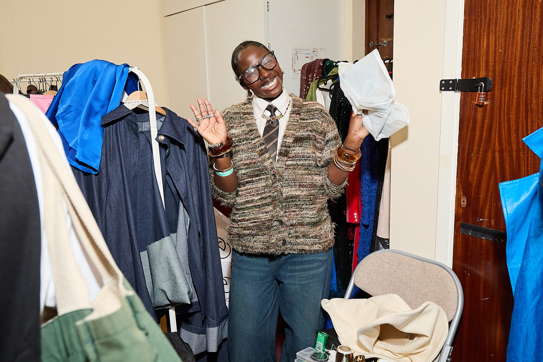 Student in behind the scenes at a busy fashion show with many garments around her smiling at the camera